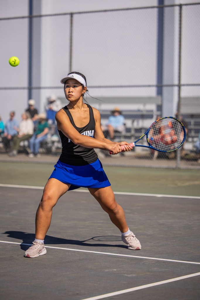 A female tennis player in a black tank top and blue skirt, preparing to hit a tennis ball with her racket on a court, surrounded by spectators in the background.