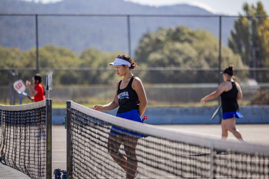 A young woman in a black tank top and blue tennis skirt is standing at the net of a tennis court, looking thoughtful. Another player in the background is preparing to hit a tennis ball.