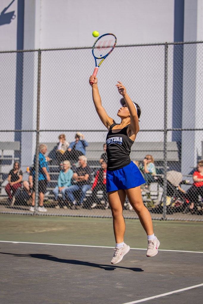 A young female tennis player in mid-air preparing to hit a yellow tennis ball with her racket on an outdoor court, with spectators in the background.