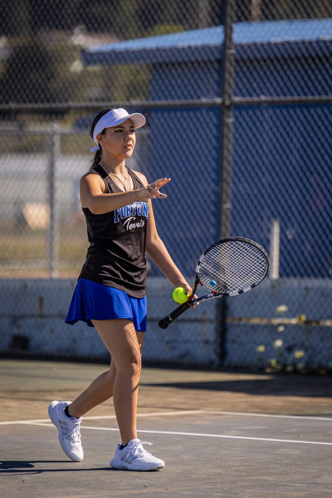 A female tennis player in a black tank top and blue skirt stands on a court, gesturing while holding a tennis racket and ball.