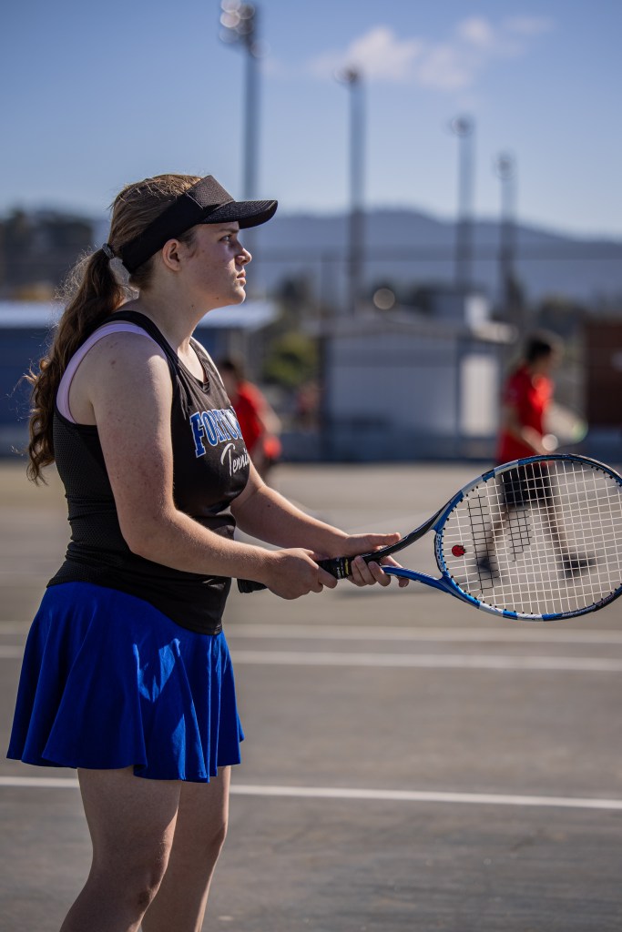 A young female tennis player in a black tank top and blue skirt, preparing to serve on an outdoor tennis court with players in the background.