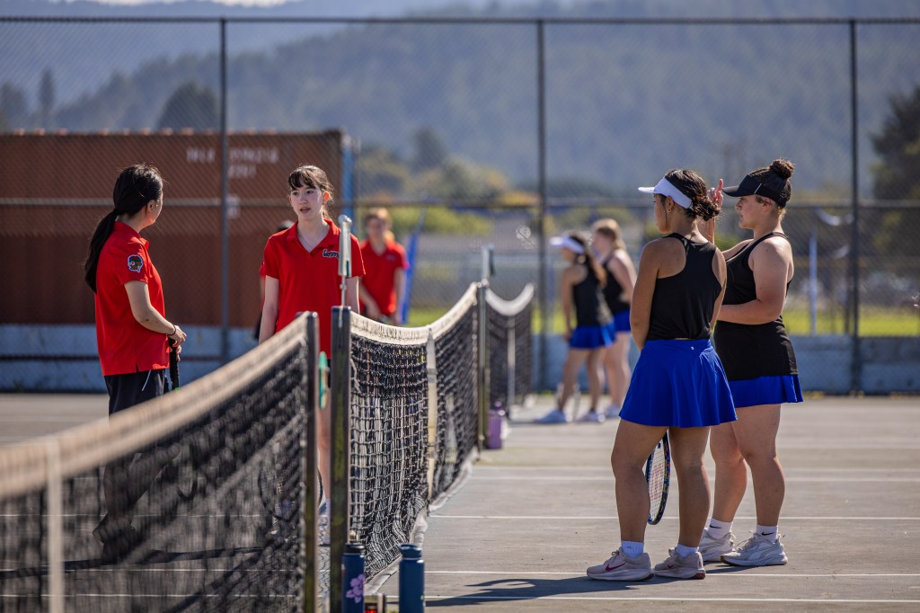 A group of young female tennis players in practice on a court, wearing colorful uniforms and interacting with coaches, with a scenic background.