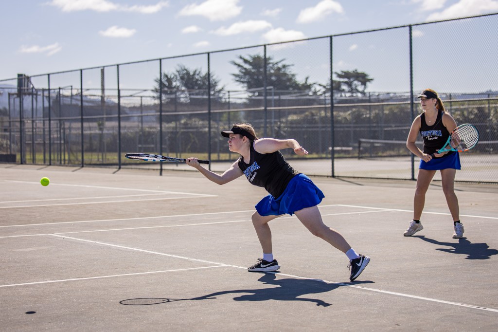 Two female tennis players on a court; one is hitting a tennis ball while the other prepares to receive.