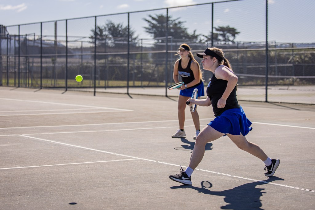 Two female tennis players on a court, one preparing to hit a ball while the other stands ready with a racquet.
