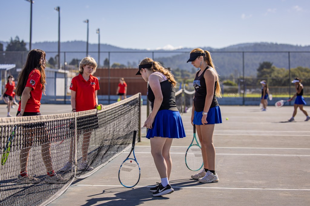 A group of female tennis players in skirts and matching tops gathered near the net on a tennis court, some practicing while others observe. The scene is set outdoors on a sunny day with hills in the background.