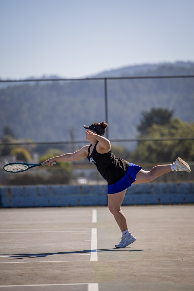 A person serves a tennis ball on a court, wearing a black tank top and blue skirt, with a tennis racket in hand.