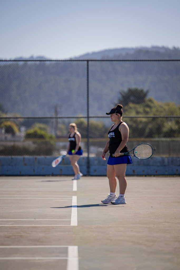 Two women playing tennis on an outdoor court, one is standing with a racket while the other is in the background preparing to serve.