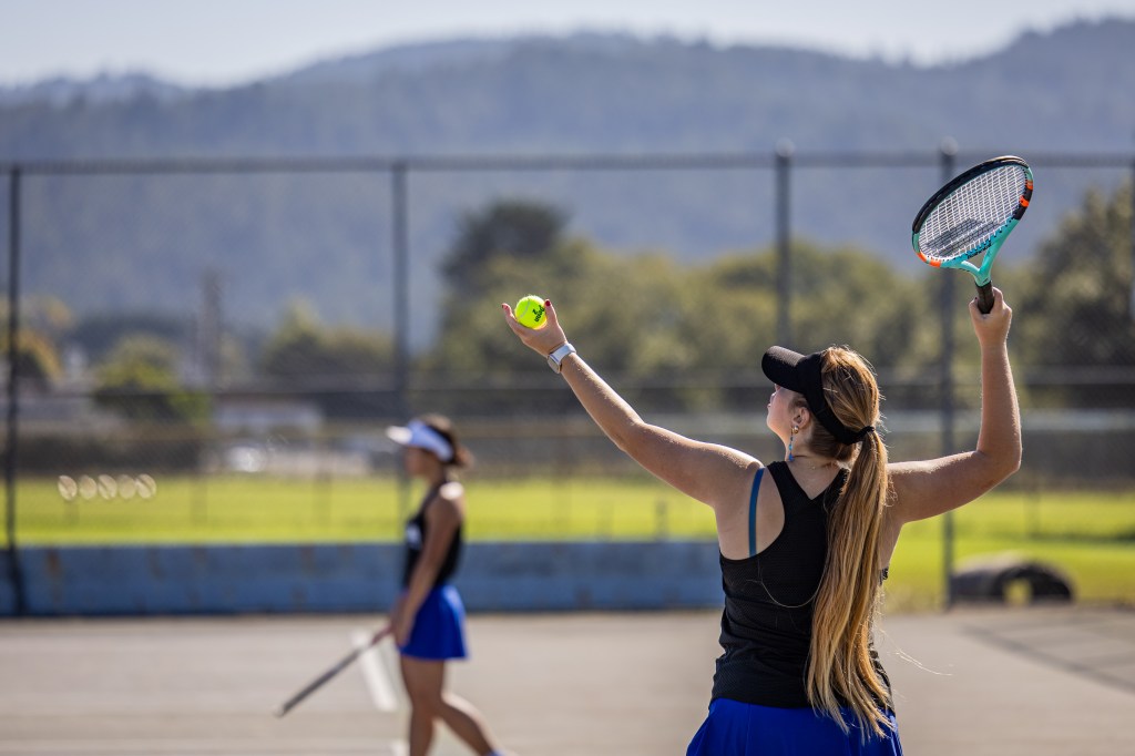 A female tennis player prepares to serve, holding a tennis ball in one hand and a racket in the other, with a second player visible in the background.