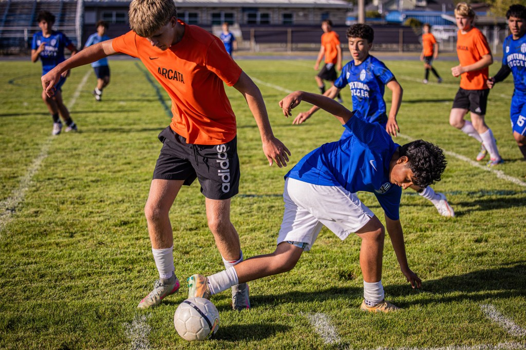 A soccer match in progress with players wearing orange and blue jerseys. One player in orange is about to intercept the ball from a player in blue who is bending down to reach it. The grassy field is marked with white lines, and other players are visible in the background.