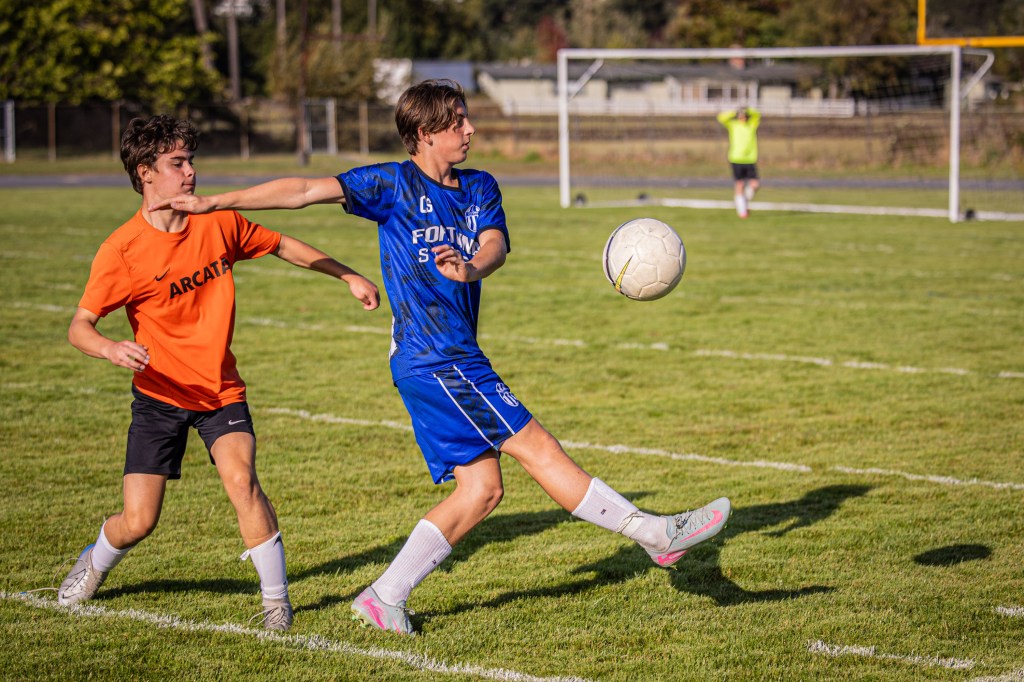 A soccer player in a blue uniform stretching to kick the ball while another player in an orange shirt reaches for him on the field.