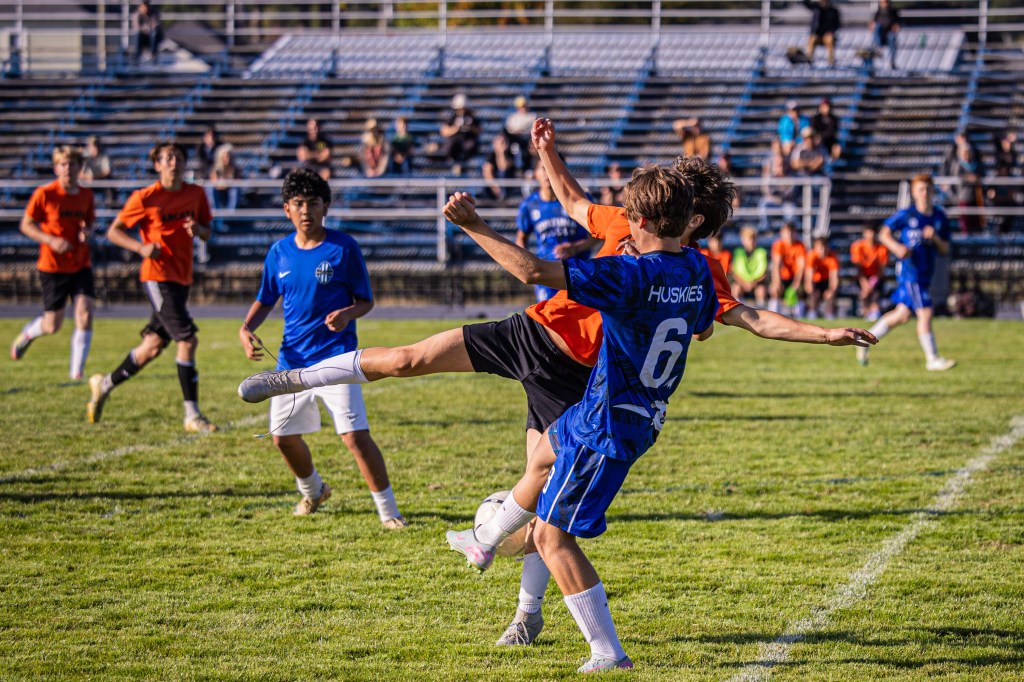 A youth soccer match featuring players in blue and orange jerseys; one player is executing a high kick while another player is attempting to block.