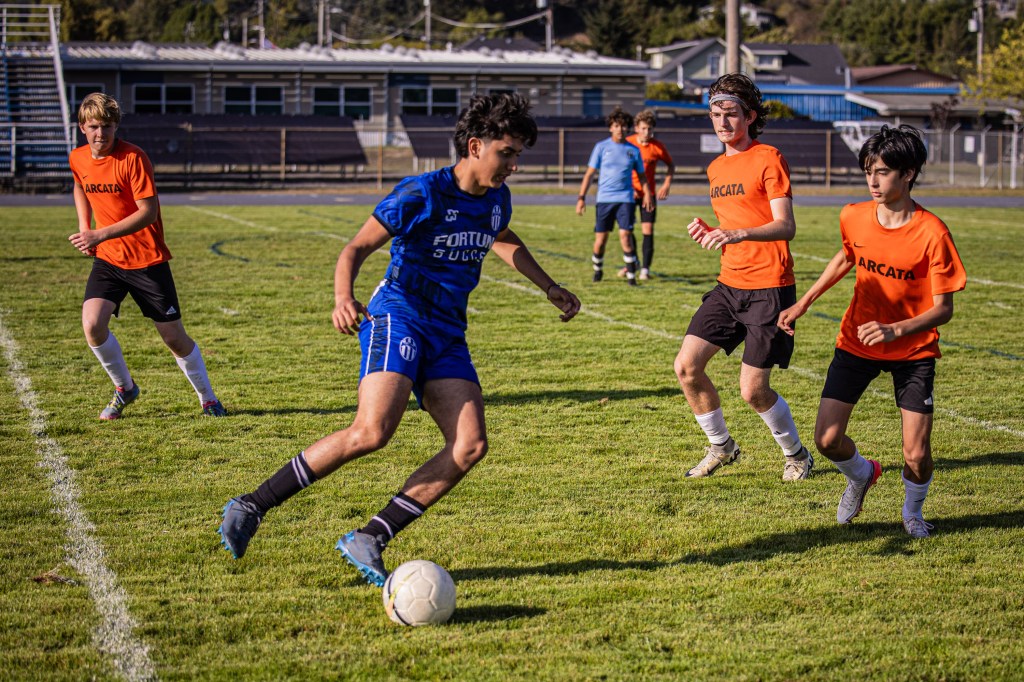 A soccer match in progress with players wearing jerseys in blue and orange. The player in blue is dribbling the ball while three players in orange and black are defending. The field is green with a goal post in the background.