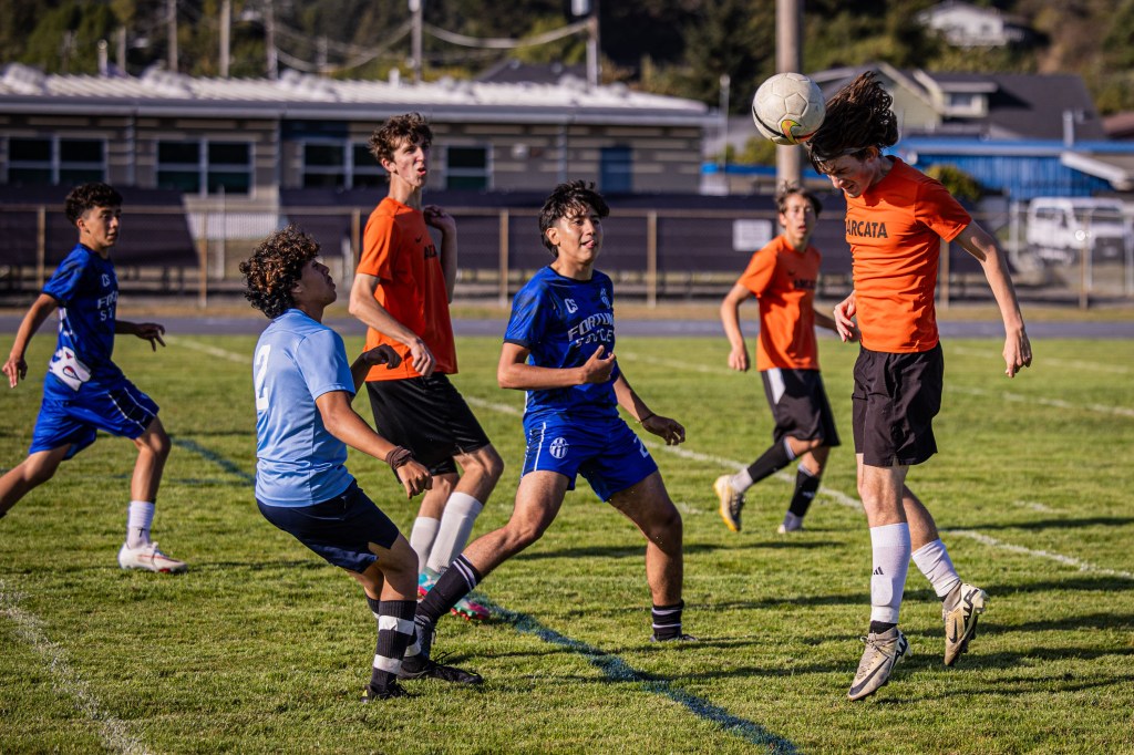 A group of young soccer players competing on a grassy field. One player in an orange shirt is jumping to head the ball, while other players in blue and black shirts are closely engaged in the play.