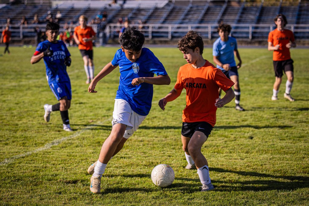 A soccer match in progress with players in blue and orange jerseys competing for the ball on a grassy field.