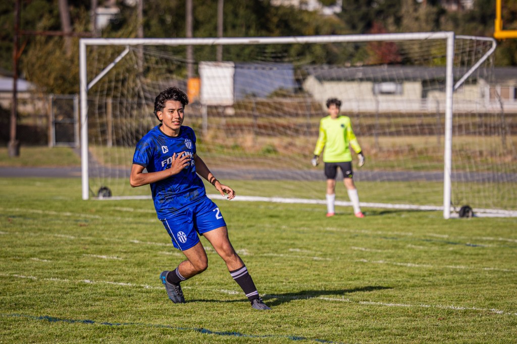 A young male soccer player wearing a blue jersey and shorts runs on a grassy field, with a goal in the background and a goalkeeper in a yellow uniform standing nearby.