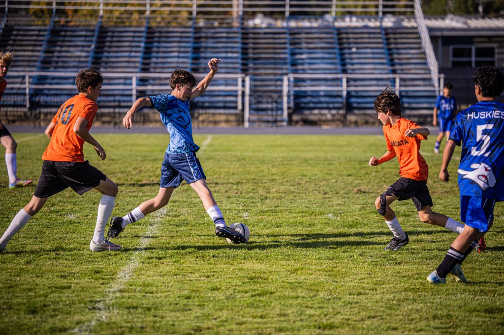 A youth soccer match in progress with players wearing orange and blue jerseys. One player in a blue jersey is about to kick the soccer ball while being surrounded by opponents in orange jerseys.