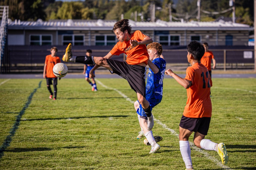 A soccer player in an orange jersey leaps into the air to kick a ball, while another player in a blue jersey attempts to defend. The scene is set on a grassy field with spectators in the background.