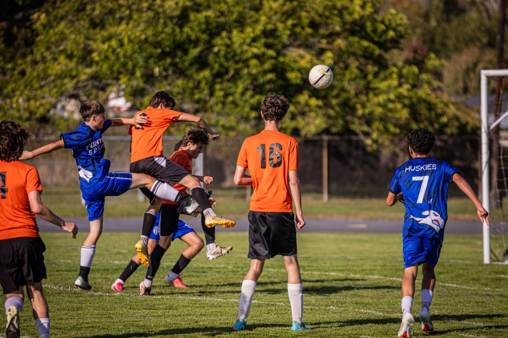 A soccer match in progress with players wearing orange and blue uniforms. One player leaps to head the ball while others attempt to defend or position themselves.