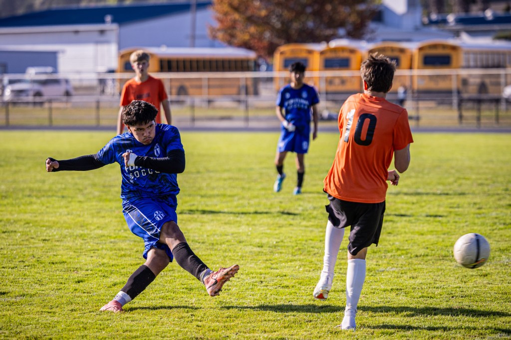 A soccer player in blue kicks the ball towards an opponent in orange during a match, with other players in the background on a grassy field.