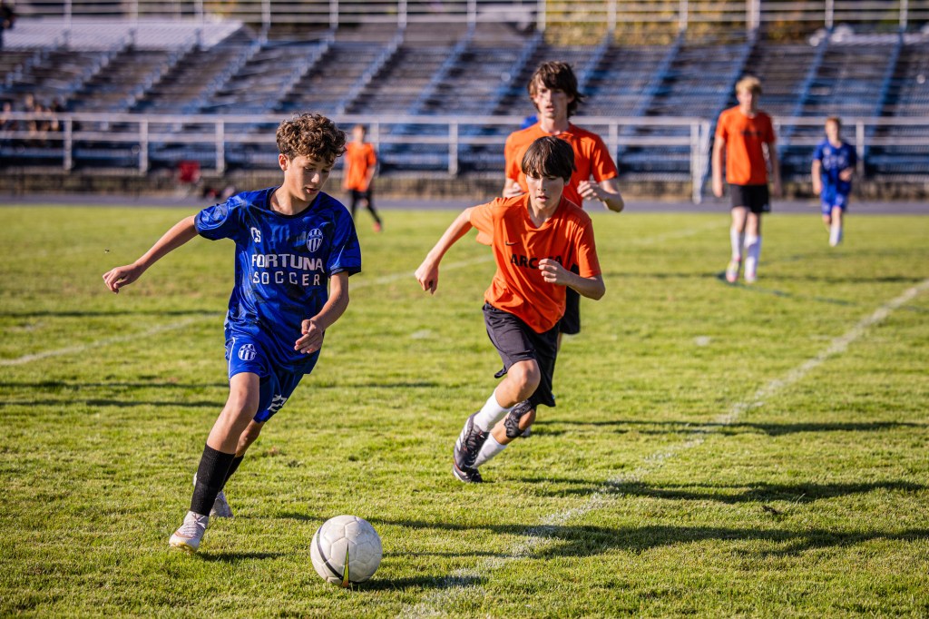 Youth soccer players running on a field during a match, with one player in a blue Fortuna jersey and others in orange uniforms.