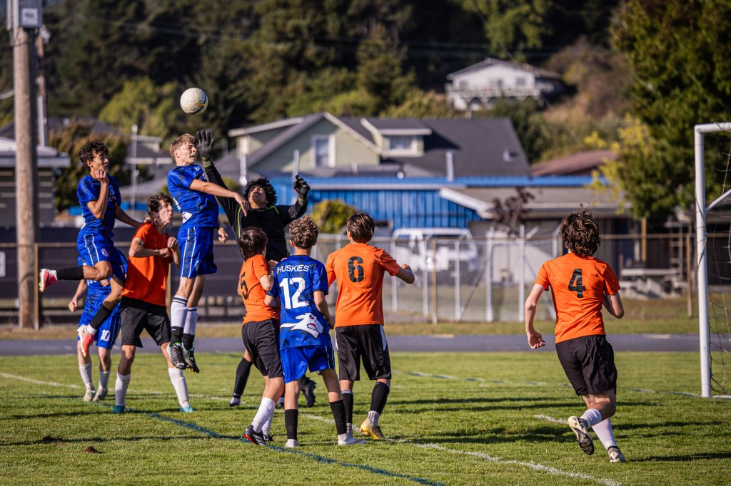 A soccer match in progress with players from two teams competing for the ball. A player in a blue jersey jumps to catch a ball while several orange jersey players attempt to block. The background features houses and trees.
