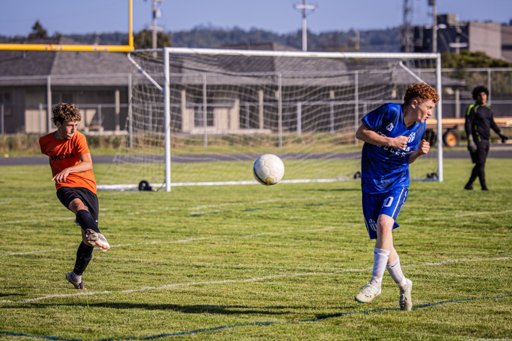 A soccer player in an orange jersey kicks the ball towards a teammate in a blue jersey, while another teammate stands nearby on the field.