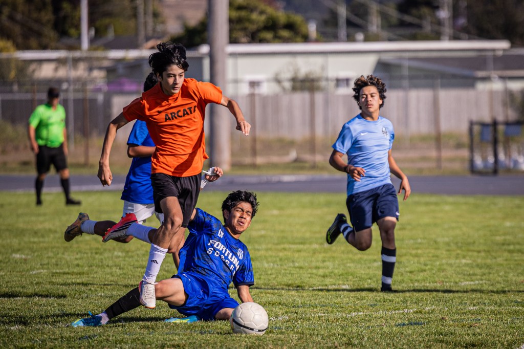 A soccer player in an orange jersey is jumping over an opponent on the ground, who is wearing a blue jersey. Two other players in blue and black jerseys are running in the background.