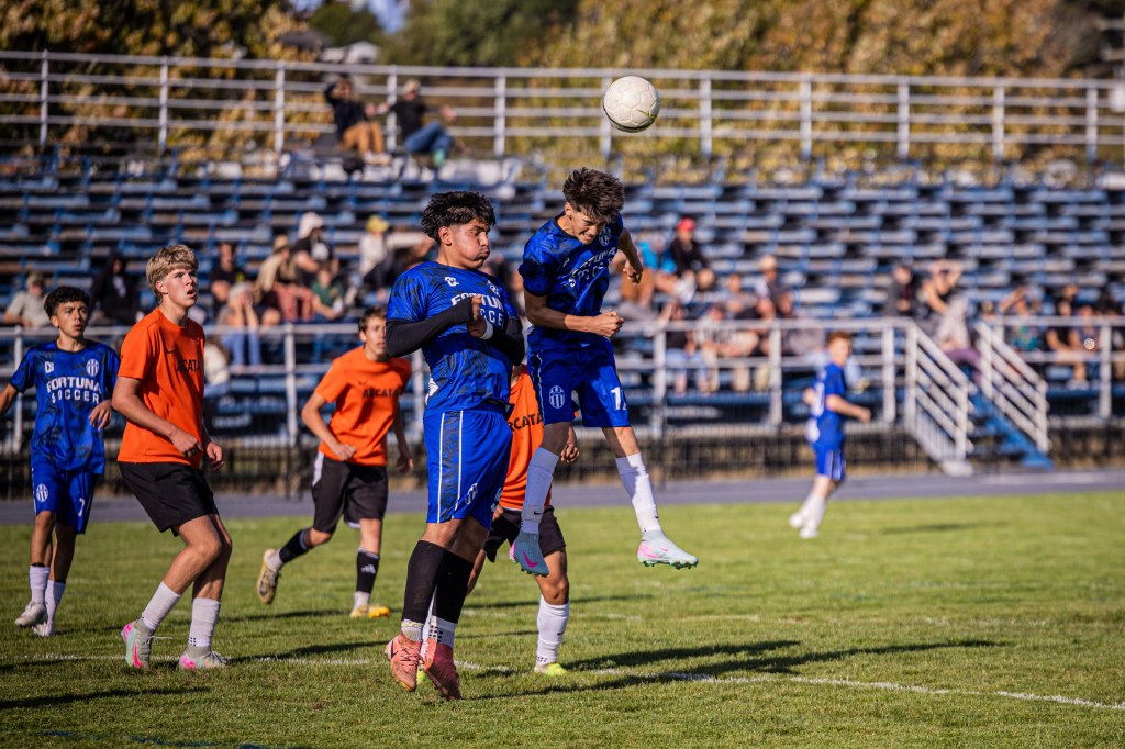 A soccer game in progress with players jumping and competing for the ball. The two main players are wearing blue jerseys with 'Fortuna Soccer' branding. The background features spectators sitting in bleachers.