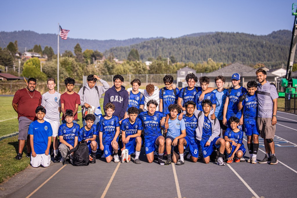 A youth soccer team poses for a group photo on a field, wearing blue jerseys with 'Fortuna Soccer' printed on them. The background features a gentle landscape with trees and hills under a clear blue sky.