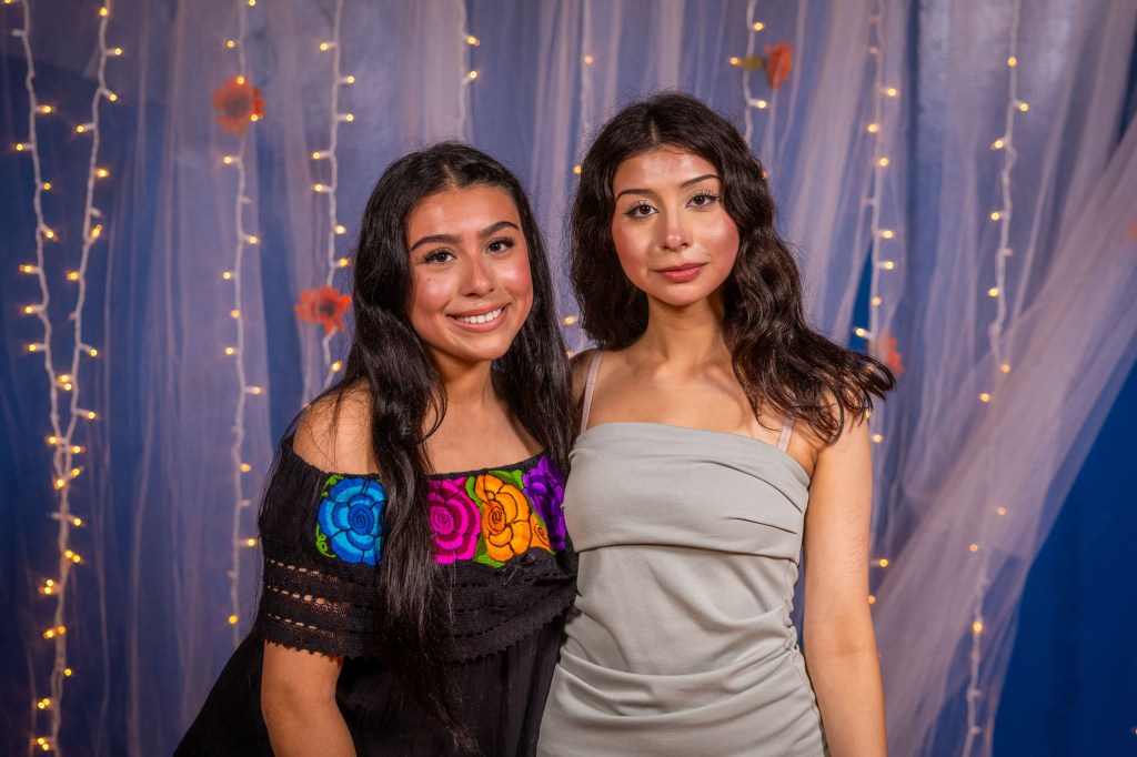 Two young women posing for a photo, one wearing a colorful embroidered blouse and the other in a sleek grey dress, with a backdrop of soft fabric and fairy lights.