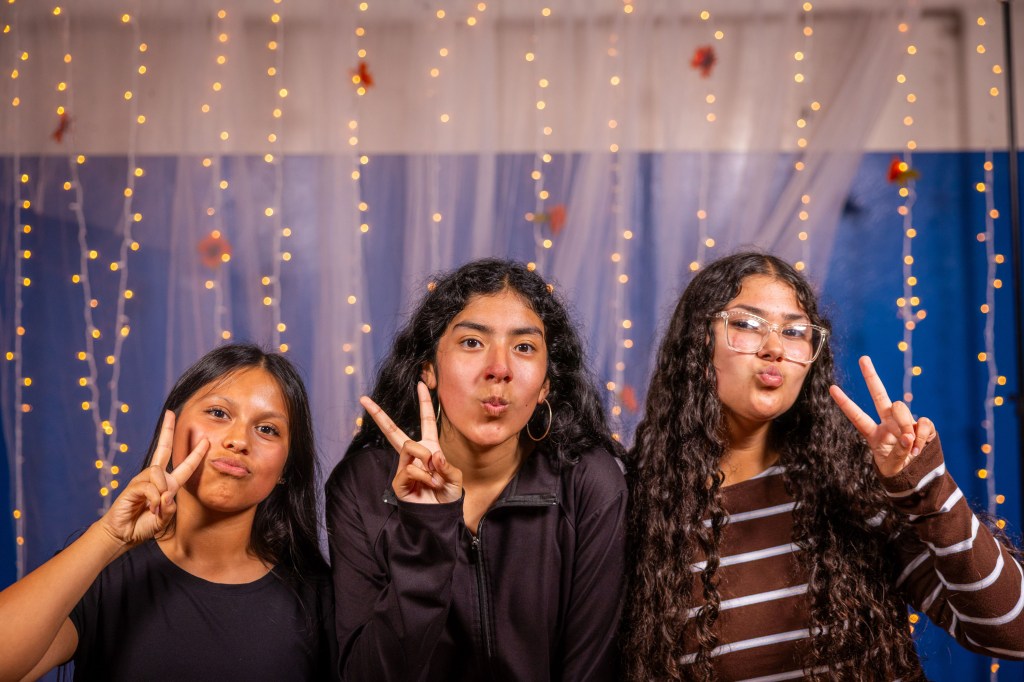 Three young girls posing playfully in front of a backdrop with fairy lights, making peace signs and playful facial expressions.