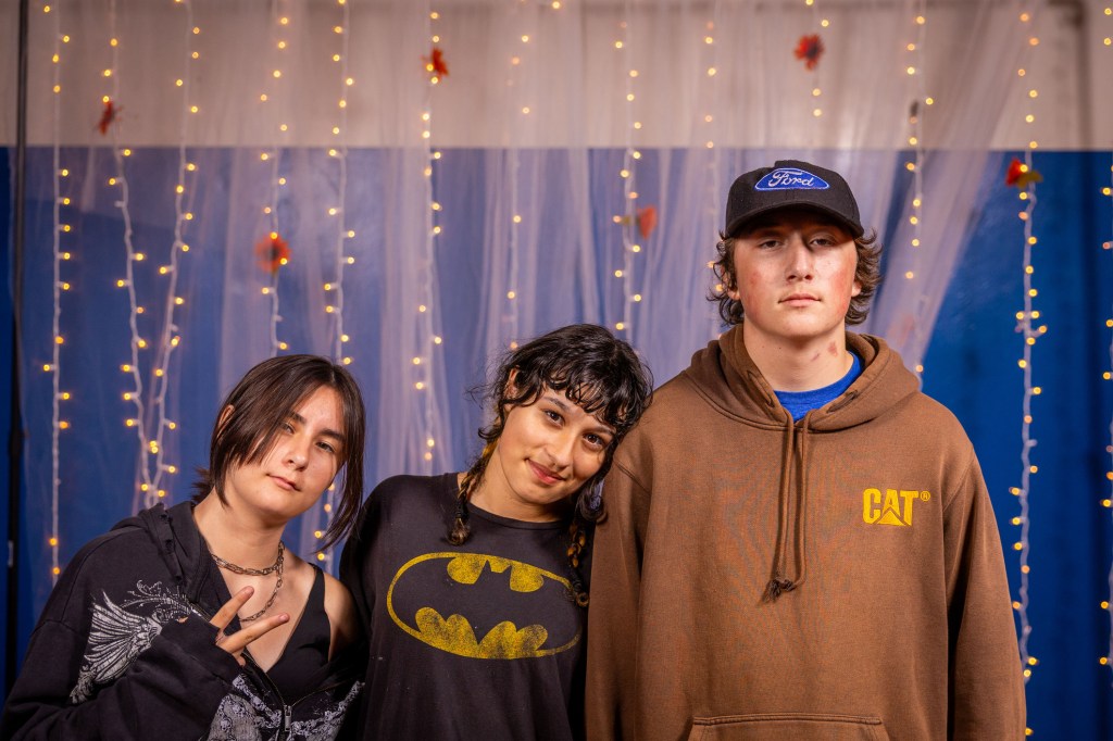 Three adolescents posing for a photo in front of a backdrop with fairy lights; one wearing a black hoodie, one in a Batman t-shirt, and the third in a brown sweatshirt.