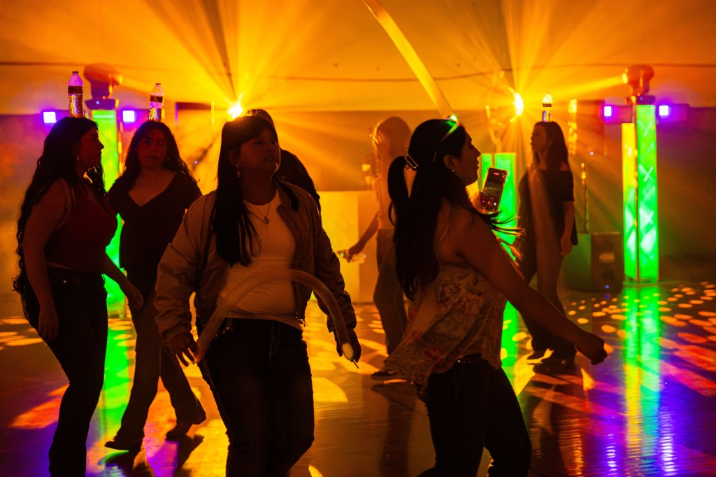 A group of women dancing joyfully under colorful lights in a lively indoor setting, with illuminated columns in the background.