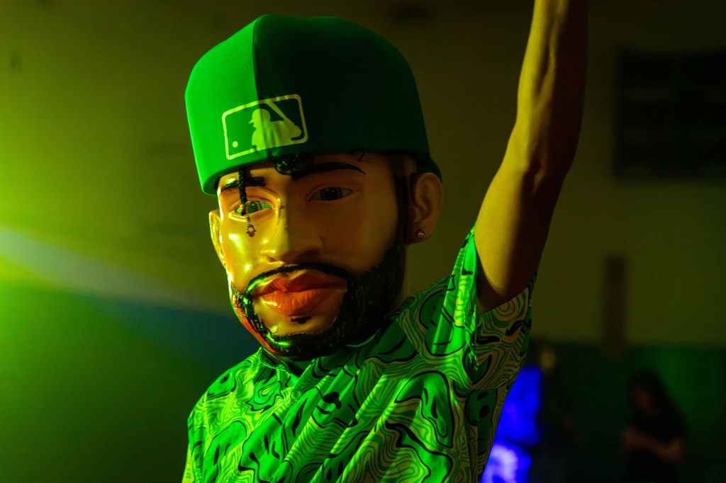 A person wearing a large, cartoonish mask with a green baseball cap, showcasing a stylized facial design, raising one arm in a lively setting illuminated by green and blue lights.