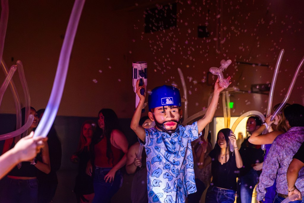 A lively party scene featuring a person wearing a large mask and holding props, surrounded by people dancing with balloon animals in a colorful, bubbly atmosphere.
