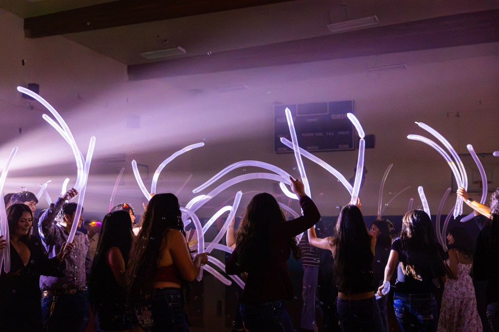 Crowd of people dancing indoors with illuminated glow sticks under colorful lighting.