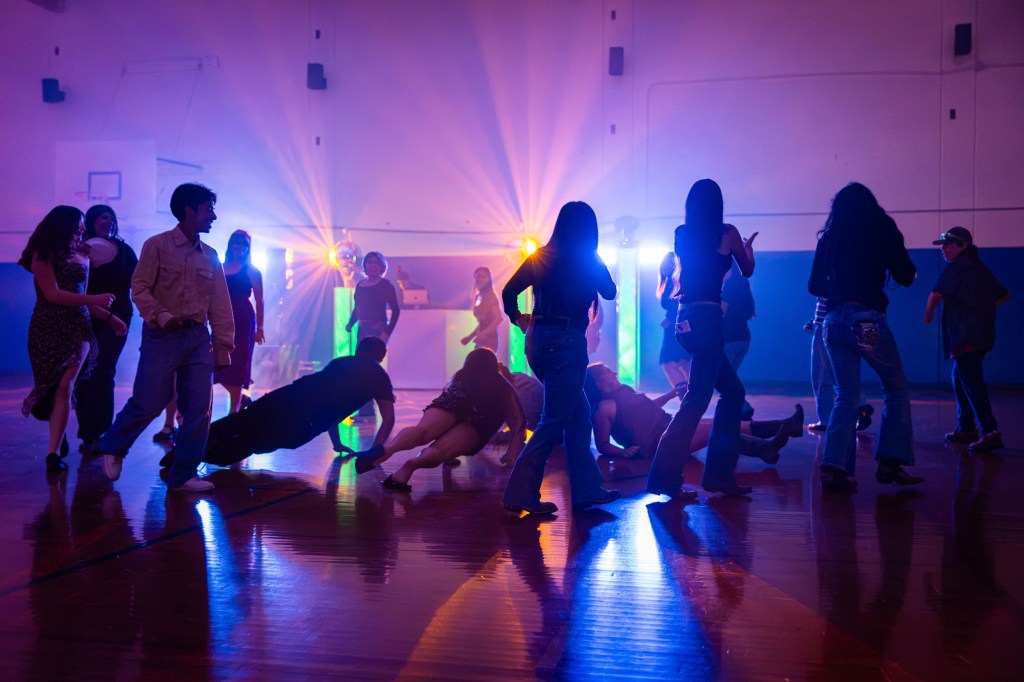 A group of people dancing in a gym, illuminated by colorful lights and a DJ setup in the background, creating a vibrant atmosphere.