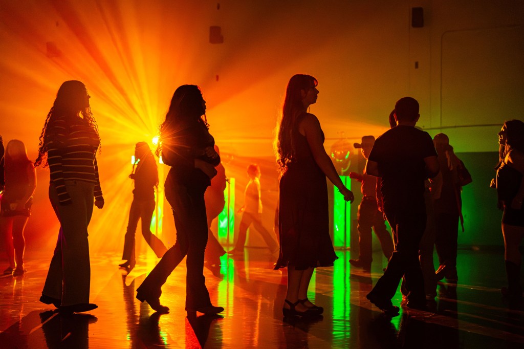 Silhouetted figures dancing under colorful lights, with vibrant orange and green beams illuminating the scene.