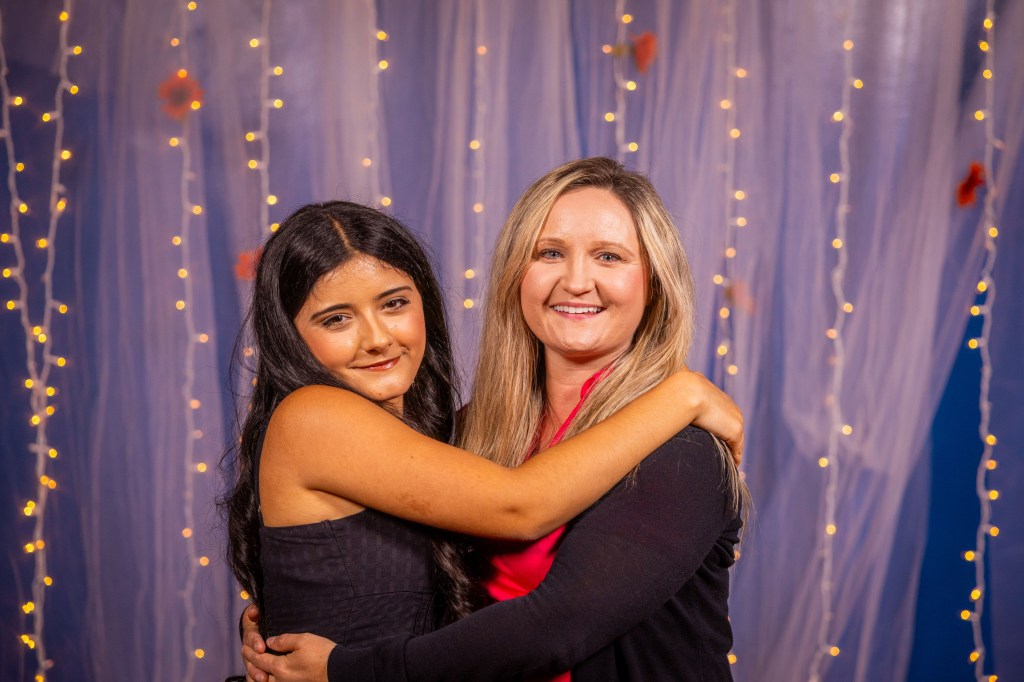 Two women smiling and embracing each other in front of a decorative background with soft lighting and draped fabric.