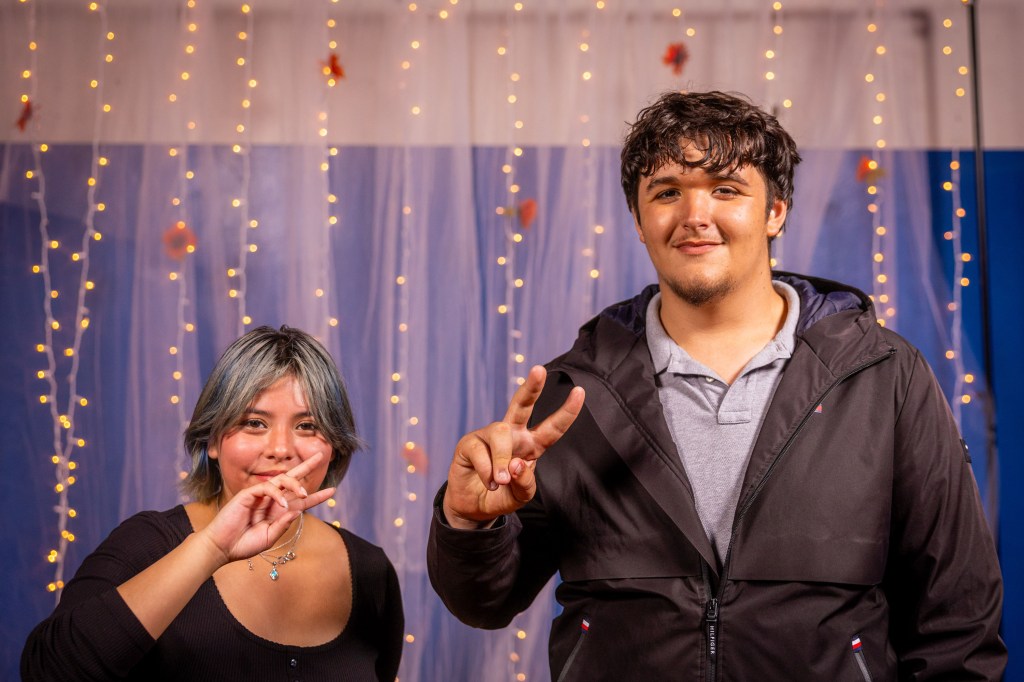 Two individuals posing for a photo, one showing a peace sign with their hand while the other holds a finger to their lips. They are in front of a decorated background with twinkling lights.