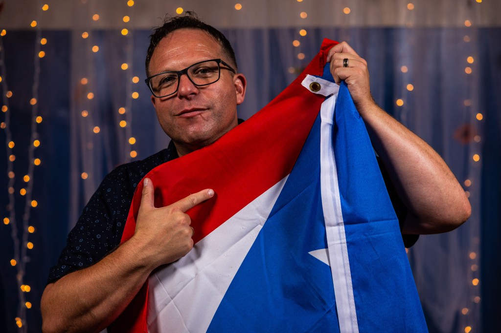 A man in glasses holds a flag featuring red, white, and blue colors while posing confidently with a playful expression.