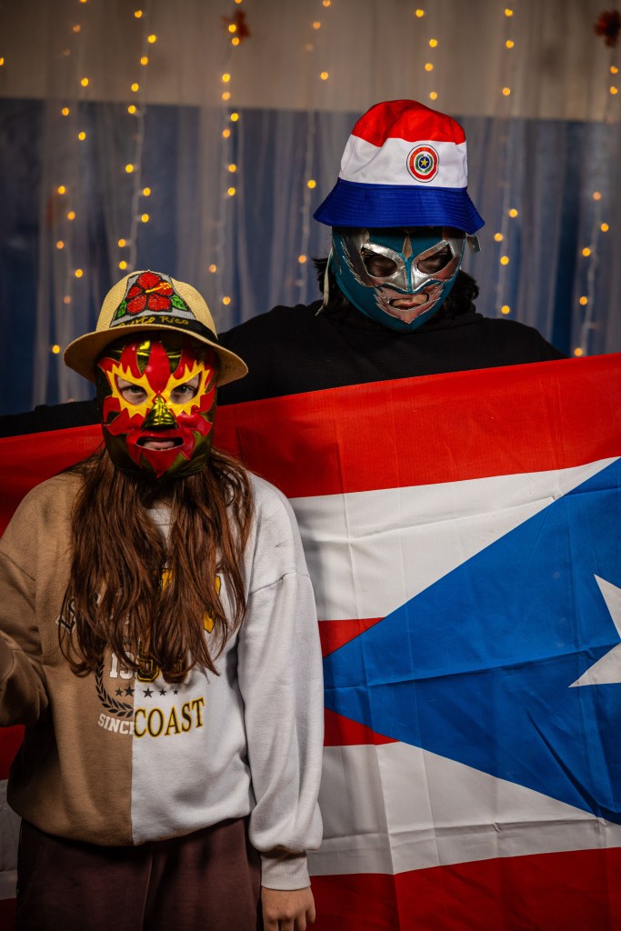 Two people wearing colorful masks and hats stand in front of a flag, one holding a flag. The background has warm string lights.