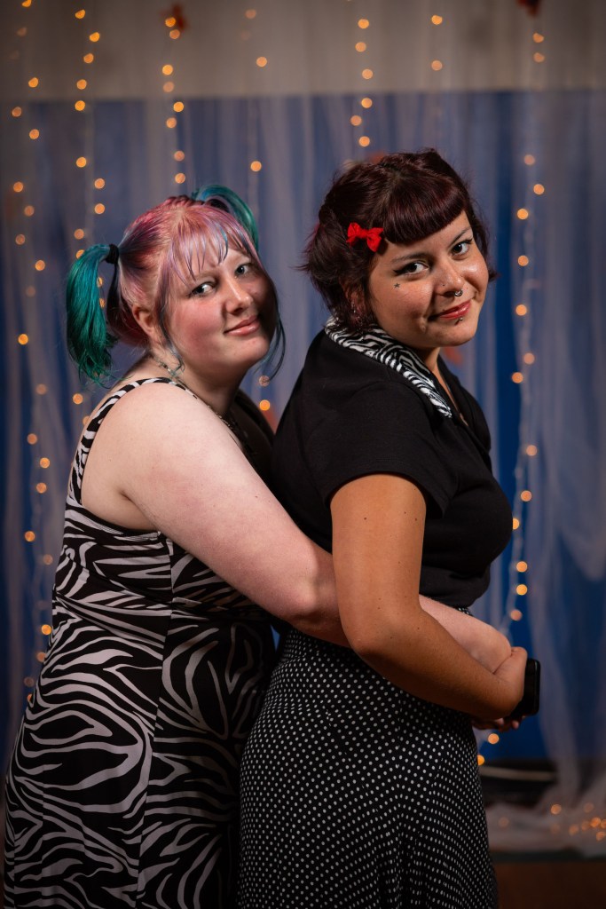 Two women posing together with one embracing the other from behind, both smiling. The woman in front has colorful hair and a zebra print dress, while the woman in back wears a dotted dress and has a red hair accessory. The background features soft lighting and decorative elements.