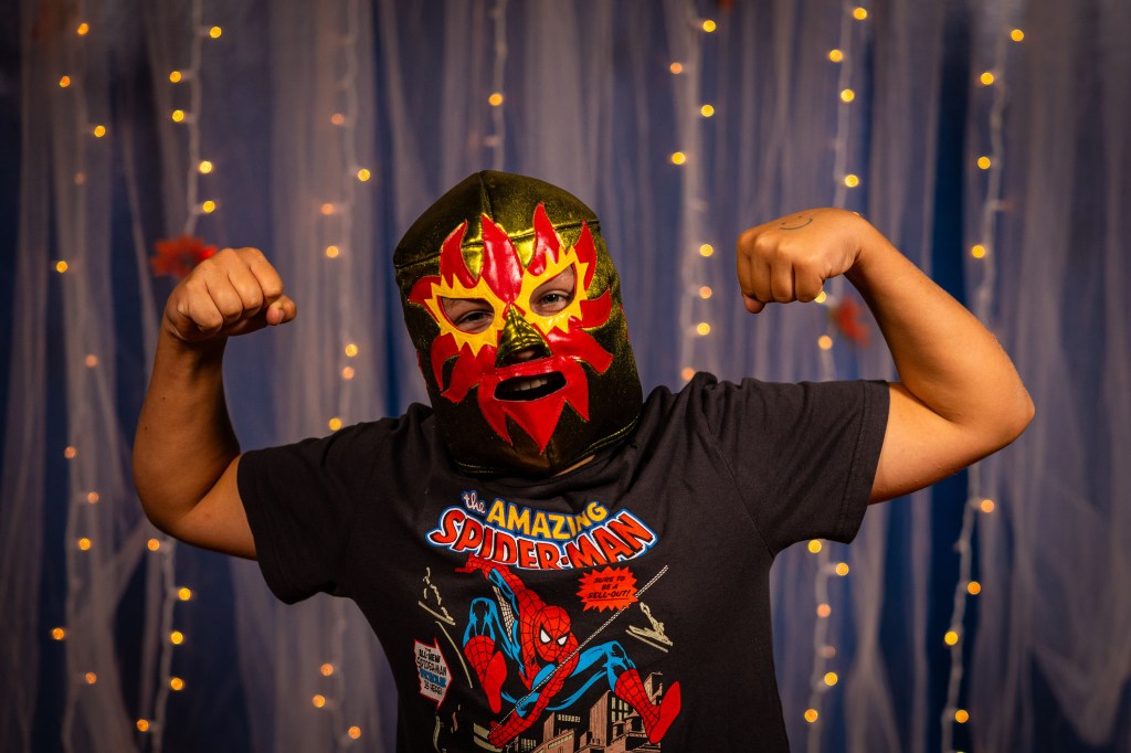 Person wearing a colorful lucha libre mask and a black 'Amazing Spider-Man' t-shirt, flexing their muscles against a backdrop with fairy lights.