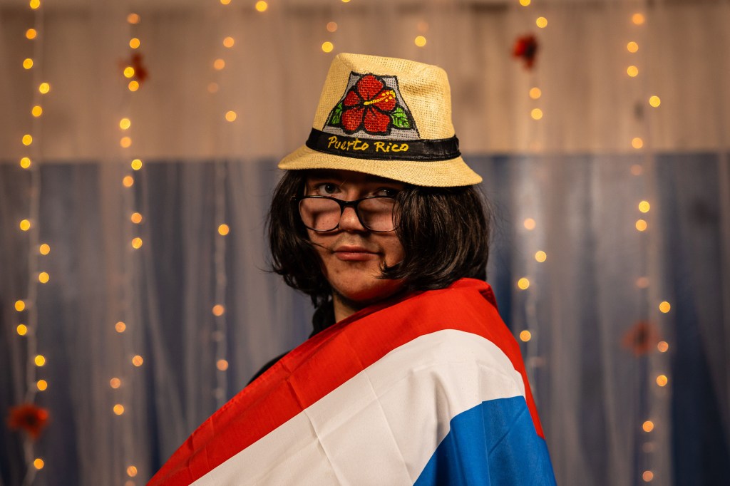 Person wearing a straw hat with 'Puerto Rico' stitched on it, draped in a Puerto Rican flag, posing against a backdrop with soft lighting.
