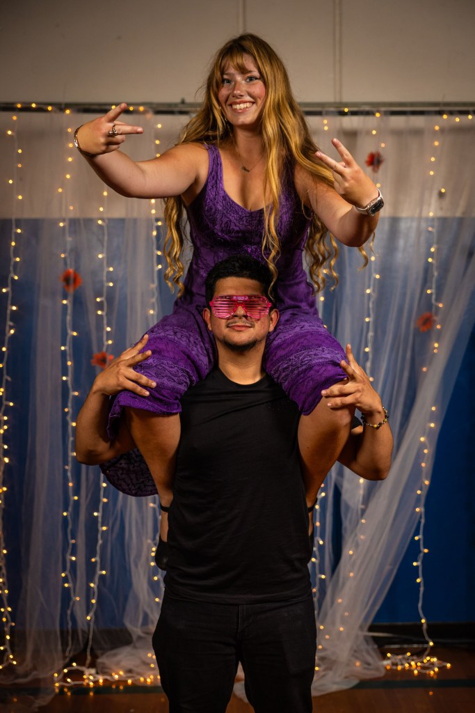 A woman in a purple dress is playfully sitting on the shoulders of a man wearing a black shirt. Both are smiling and posing with peace signs in front of a backdrop with fairy lights.