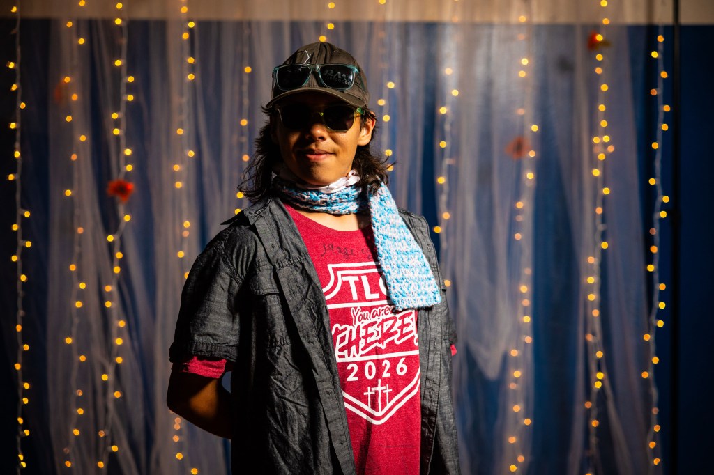 A young person wearing sunglasses and a cap, dressed in a red shirt and a light blue scarf, stands confidently in front of a background adorned with soft fairy lights and sheer drapery.
