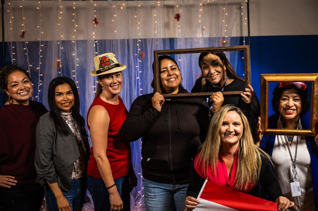 A group of seven women posing together for a photo, some holding frames and a flag, with a festive background featuring fairy lights.