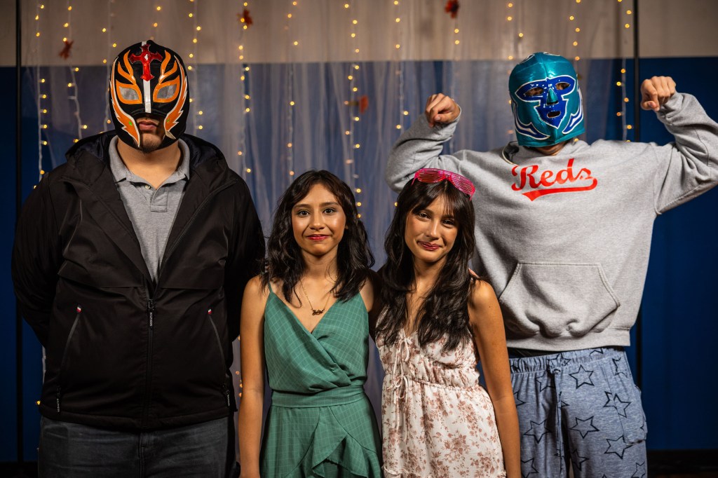 A group of four people posing together, two wearing colorful wrestling masks, with a festive backdrop of fairy lights. The individuals include two women in dresses and two men, one in a hoodie and the other in a jacket.