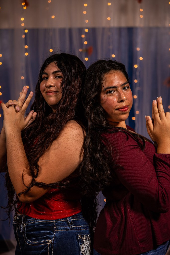 Two women posing back-to-back with playful expressions, each making finger gun gestures. The background features soft lighting and a colorful backdrop.